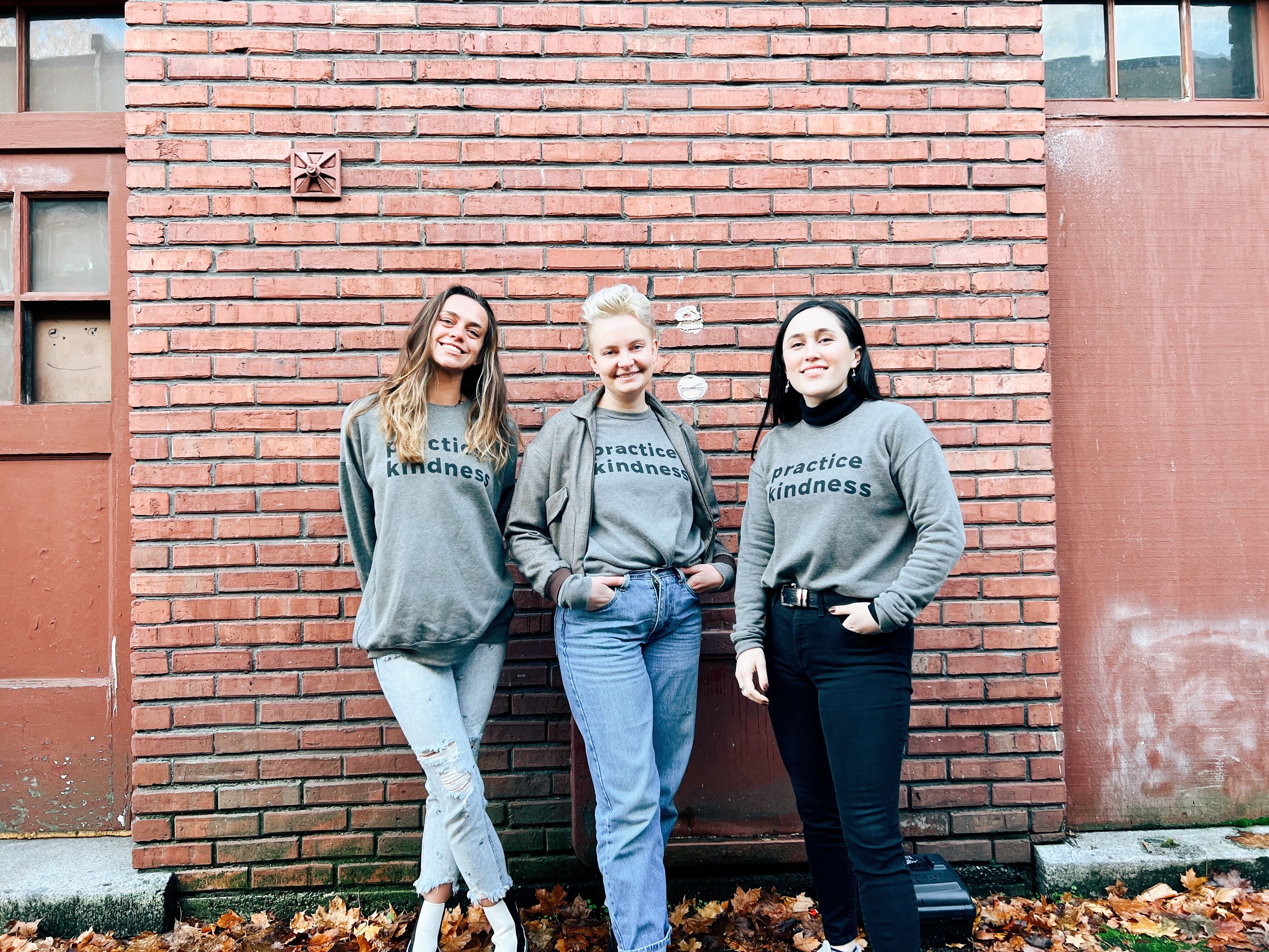 3 people stand with gray sweatshirts on that say Practice Kindness in dark gray bold font.