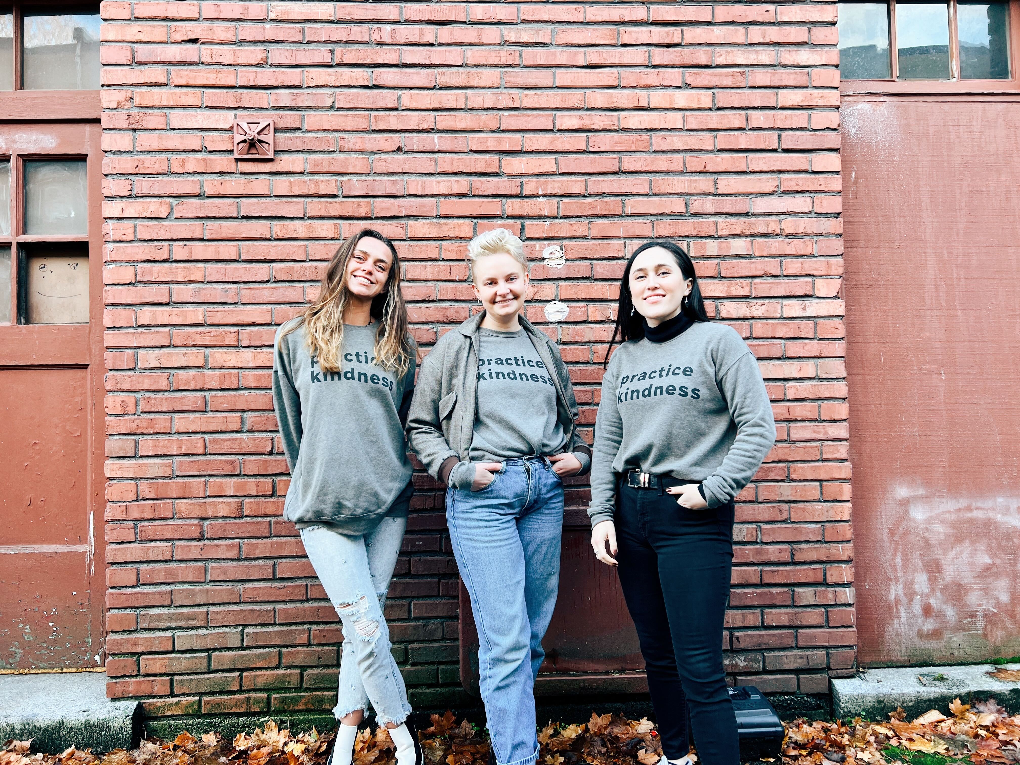 Sweatshirts 1 3 people stand with gray sweatshirts on that say Practice Kindness in dark gray bold font.